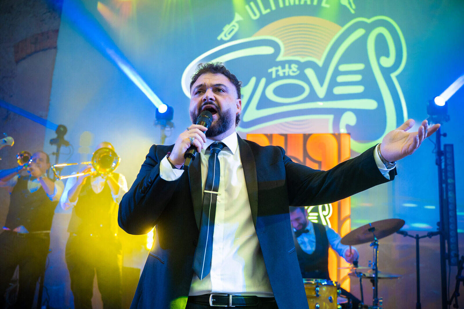 Il cantante dei The Gloves al microfono con un braccio aperto, fiati e batteria sullo sfondo e luci di scena blu e gialle.