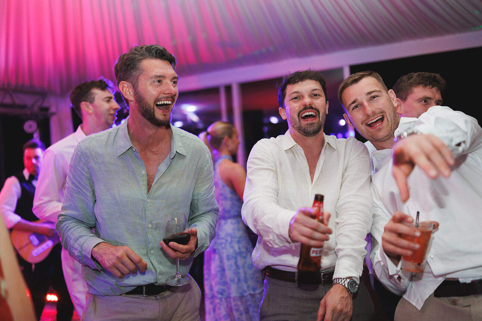 Tre invitati sorridenti con bicchieri in mano durante il party, sotto le luci rosa del padiglione.