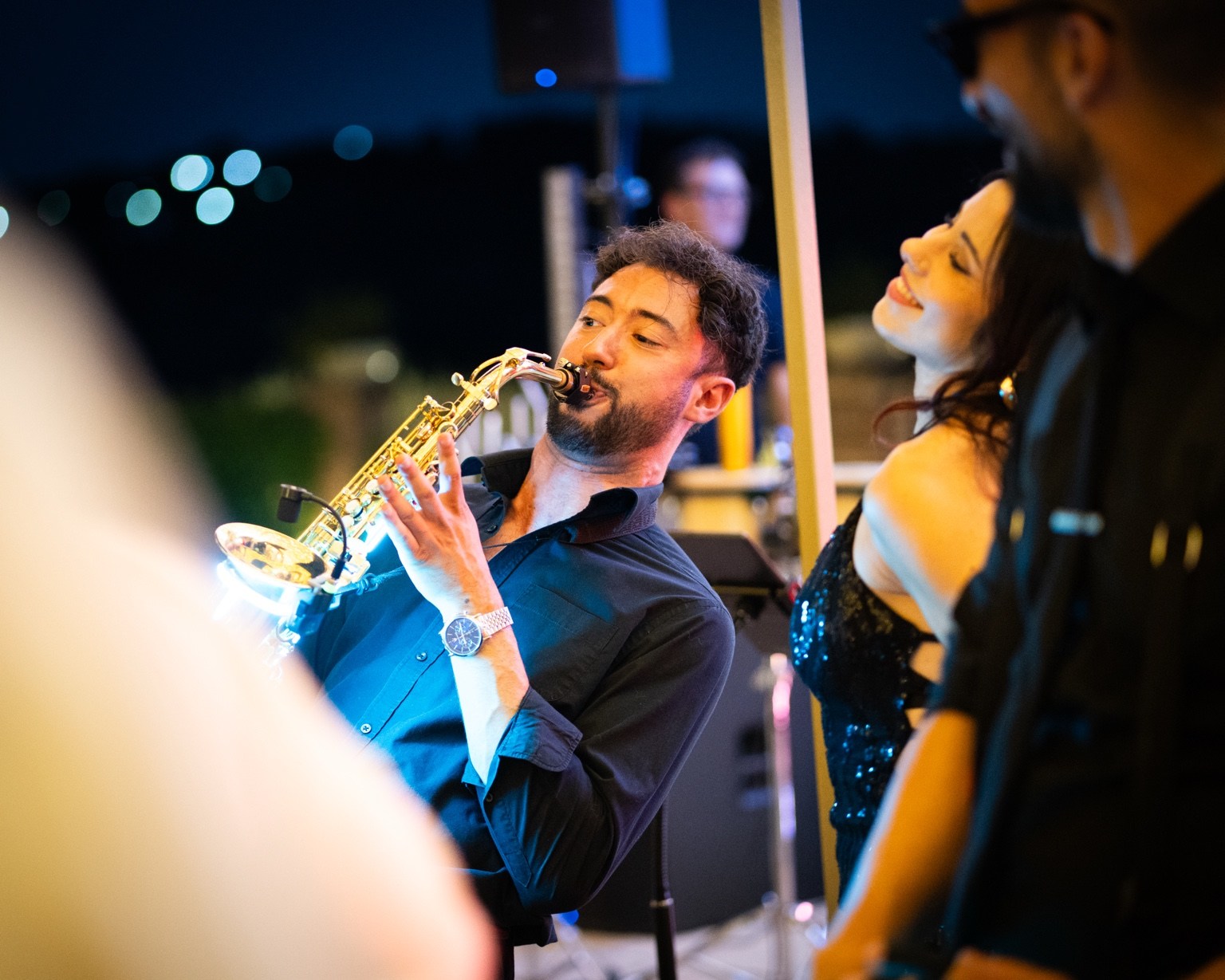 Saxophonist performing beside singers under coloured wedding party lights.