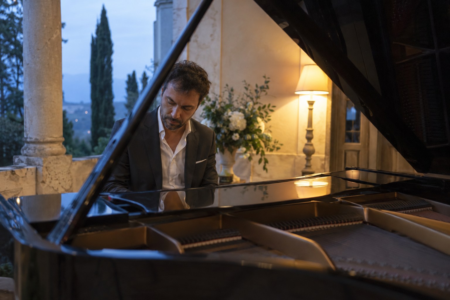 Pianist playing a black grand piano in a villa loggia at dusk.