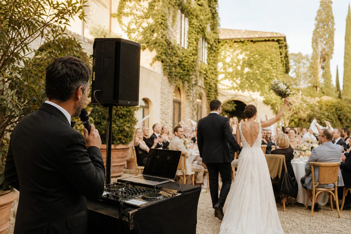 DJ holding a microphone beside a mixer, laptop and speaker as newlyweds enter an outdoor dinner reception.