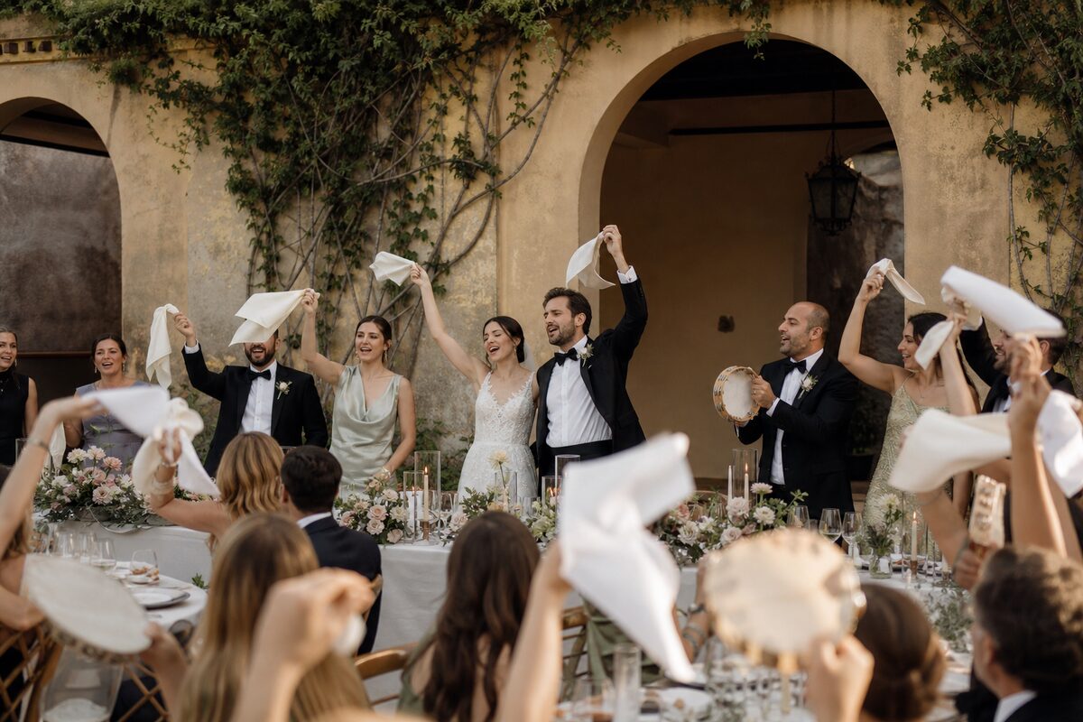 Newlyweds and bridal party standing behind a decorated head table as guests wave napkins and tambourines.