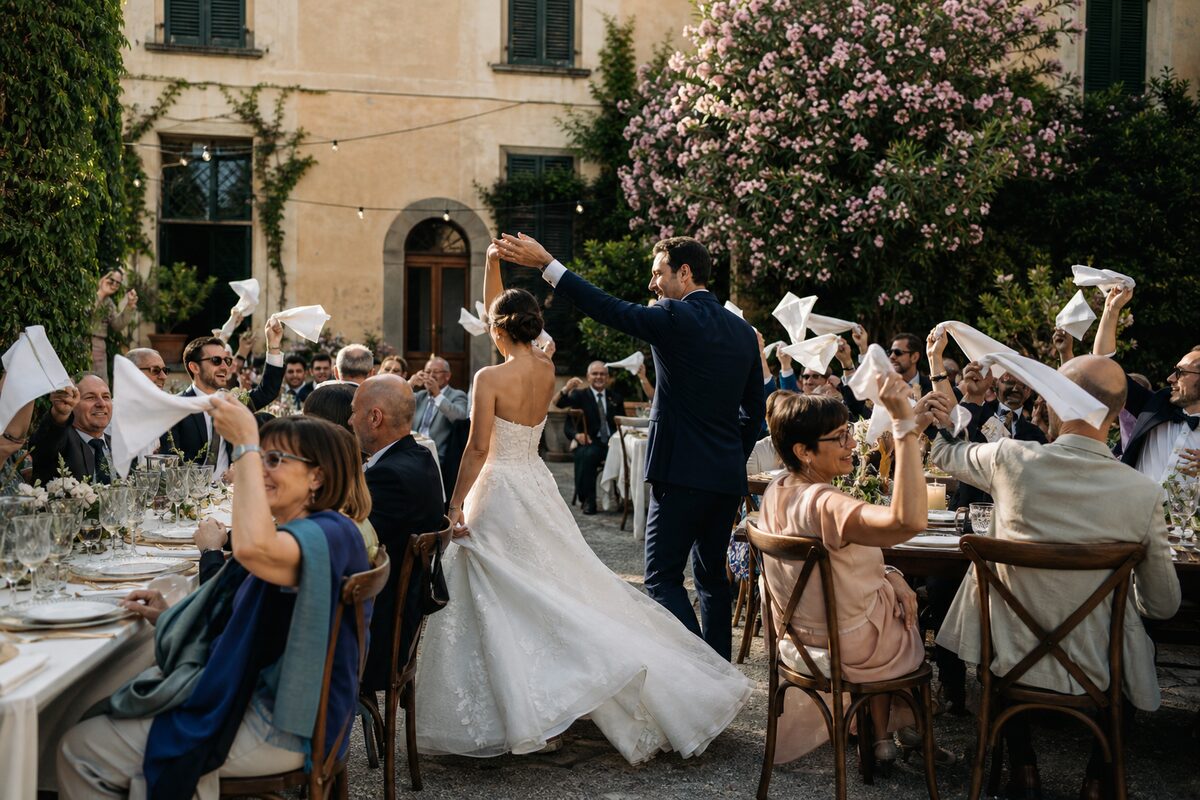 Newlywed couple dancing through an outdoor reception as seated guests wave white napkins.