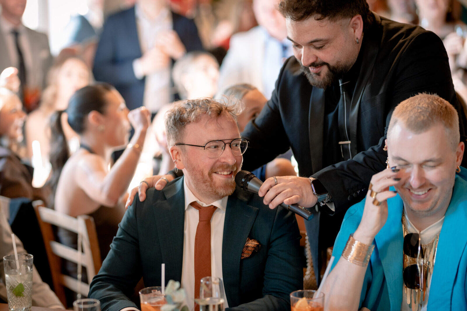 A man holds a microphone to a seated guest while the man beside him smiles with a phone to his ear.