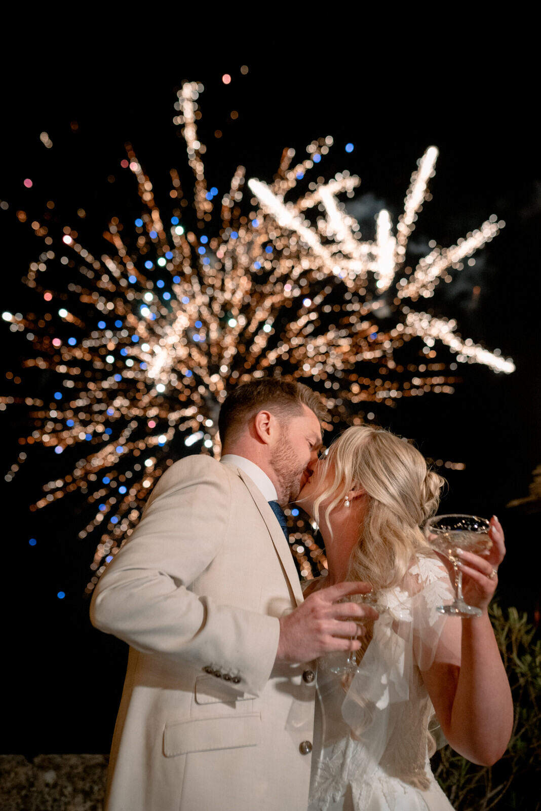 The couple kiss while holding glasses, with fireworks lighting the night sky behind them.
