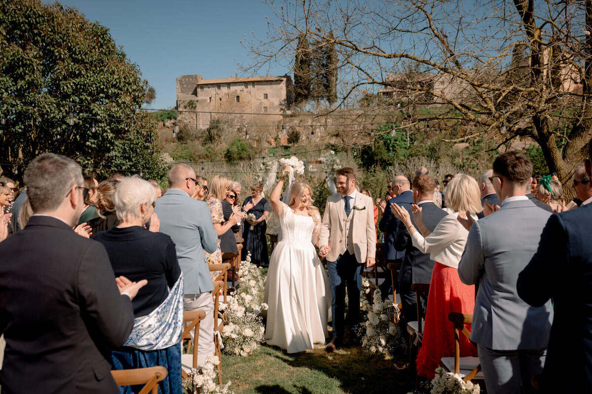 The couple walk down the outdoor ceremony aisle between standing guests, with the bride holding her bouquet aloft.