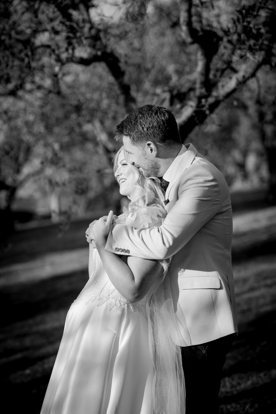 Black-and-white portrait of the groom embracing the bride from behind and kissing her head.