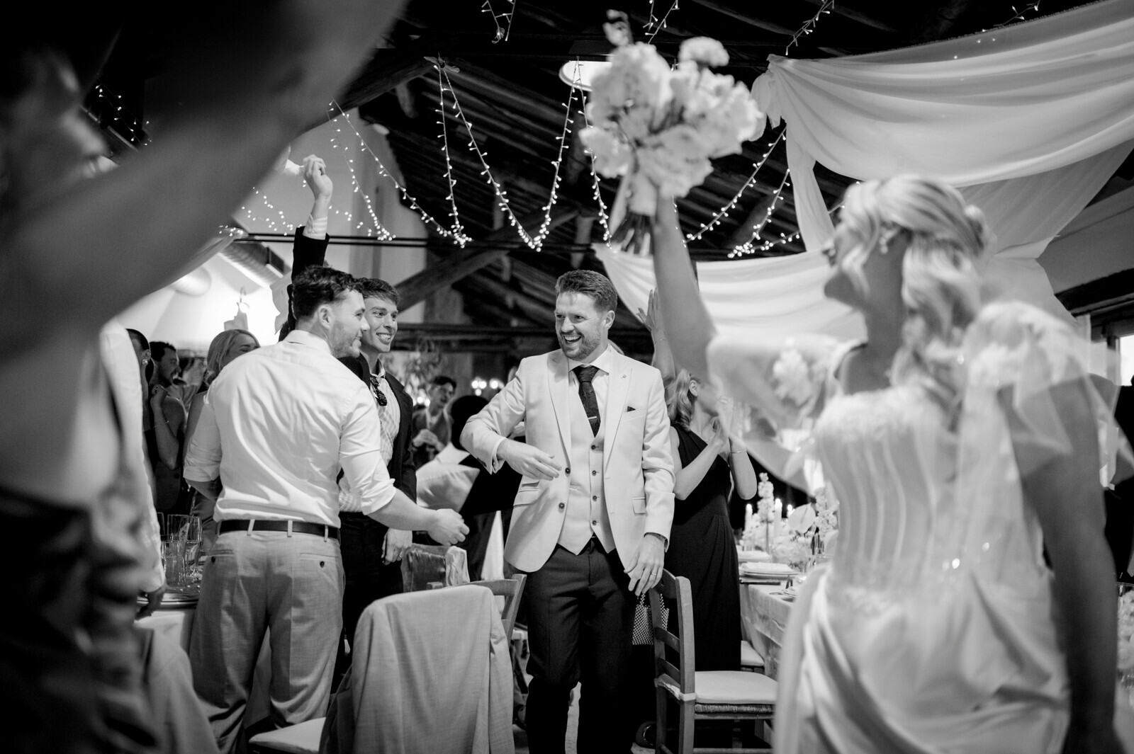 Black-and-white photograph of the couple among reception tables; the bride raises her bouquet beneath strings of lights.