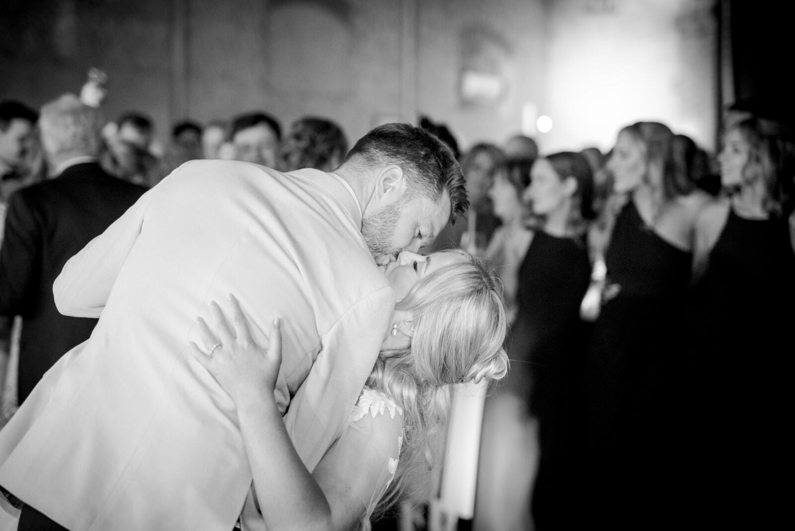 Black-and-white first-dance photograph: the groom bends towards the bride, with guests gathered behind them.