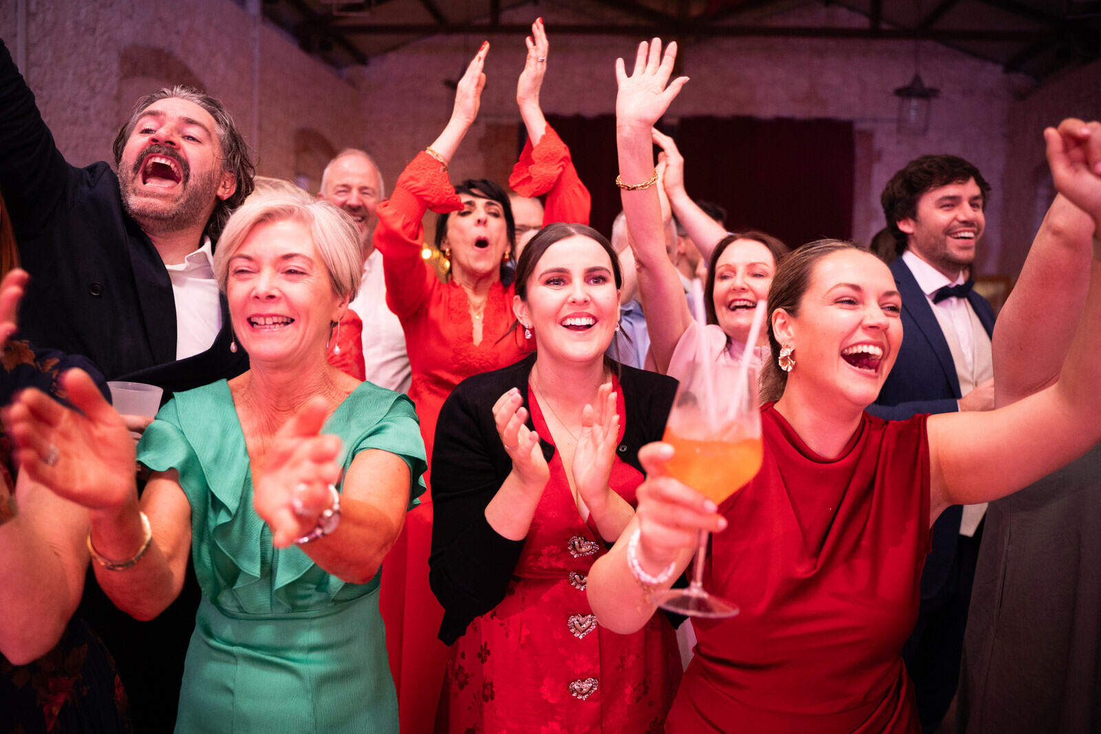 Gli invitati sorridono, applaudono e alzano le braccia durante la festa; una donna in primo piano tiene un calice.