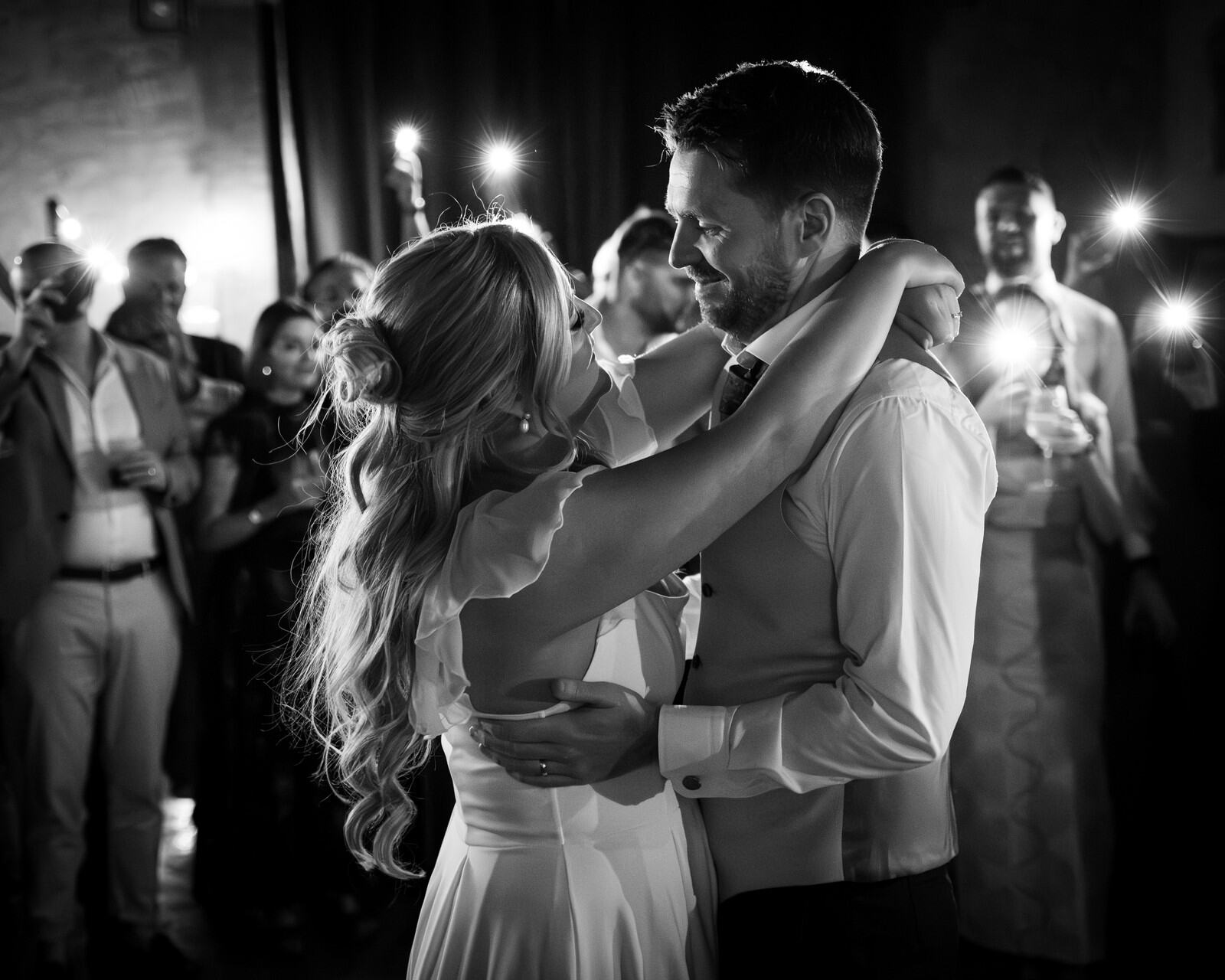 Gli sposi si abbracciano durante un ballo, circondati dagli invitati; fotografia in bianco e nero con luci sullo sfondo.