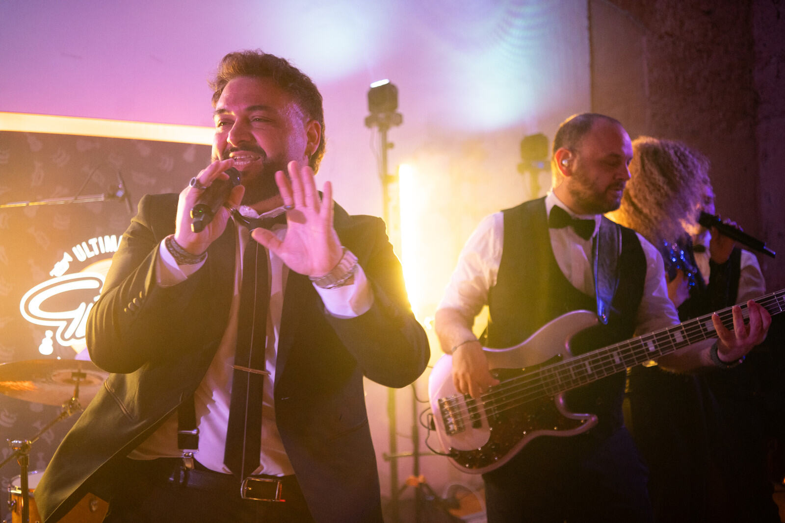 Un cantante sorride al microfono con una mano alzata, affiancato da un bassista tra luci rosa e gialle.