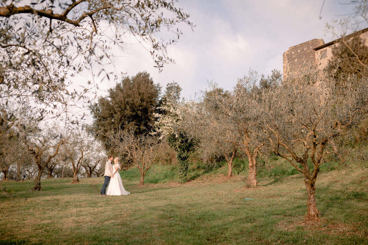 The couple stand on the lawn among olive trees at Borgo di Tragliata, with stone buildings behind them.