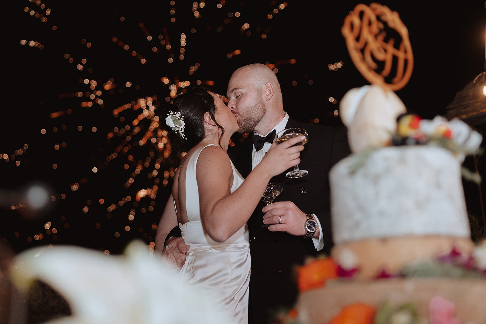 Rebecca e Hugh si baciano accanto alla torta nuziale, con fontane luminose sullo sfondo.