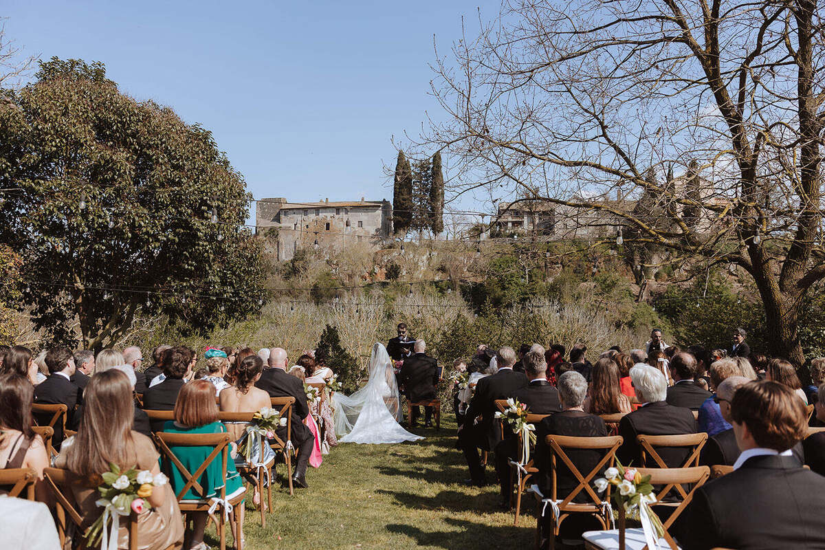 Cerimonia all’aperto di Rebecca e Hugh, con invitati seduti in giardino e il Borgo sullo sfondo.