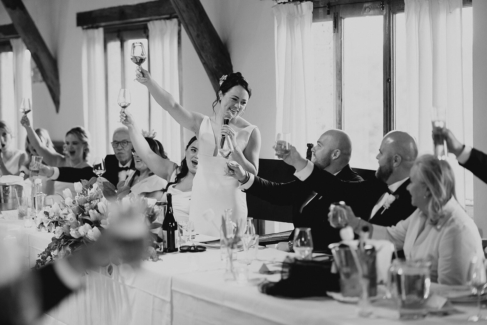 Foto in bianco e nero di Rebecca che alza il calice al tavolo del ricevimento, accanto a Hugh.