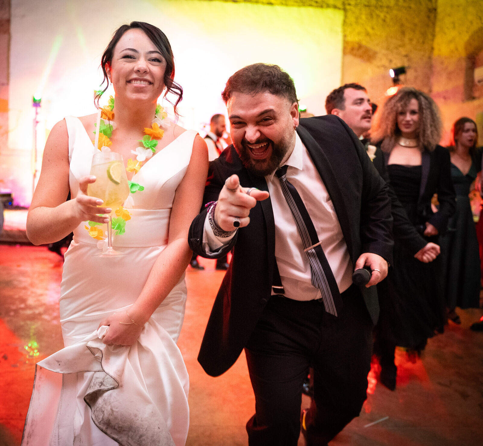 Rebecca sorride con il bouquet accanto a un invitato che indica l’obiettivo durante la festa.