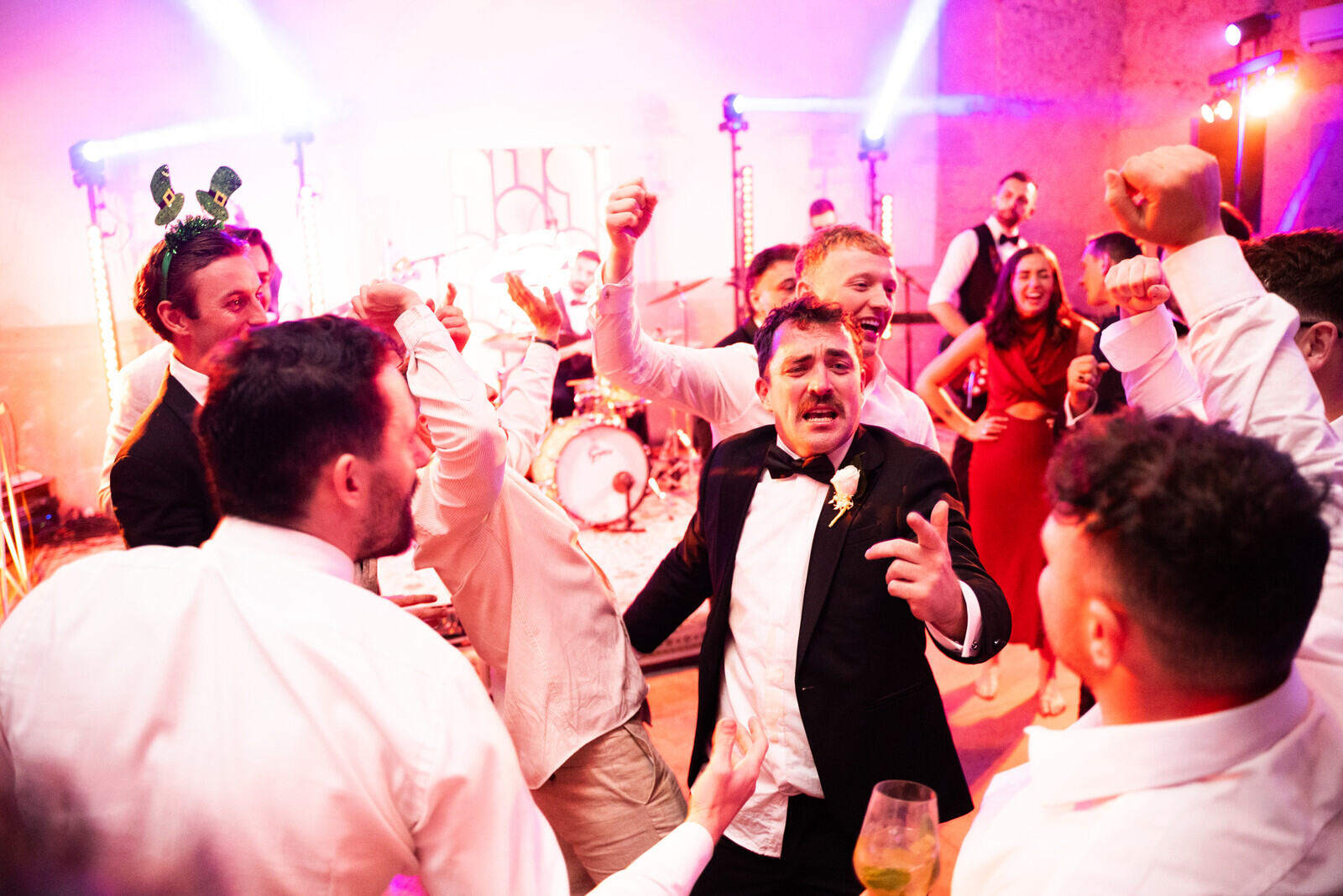 Invitati ballano con le braccia alzate davanti alla band, sotto luci rosa e viola.
